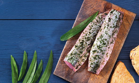 Frische Lachsfilets mit Kräutern auf Holzbrett Zwei Lachsfilets, gewürzt mit Kräutern, liegen auf einem hölzernen Schneidebrett, umgeben von frischen grünen Blättern und einer Scheibe Brot auf einem blauen Holztisch.
