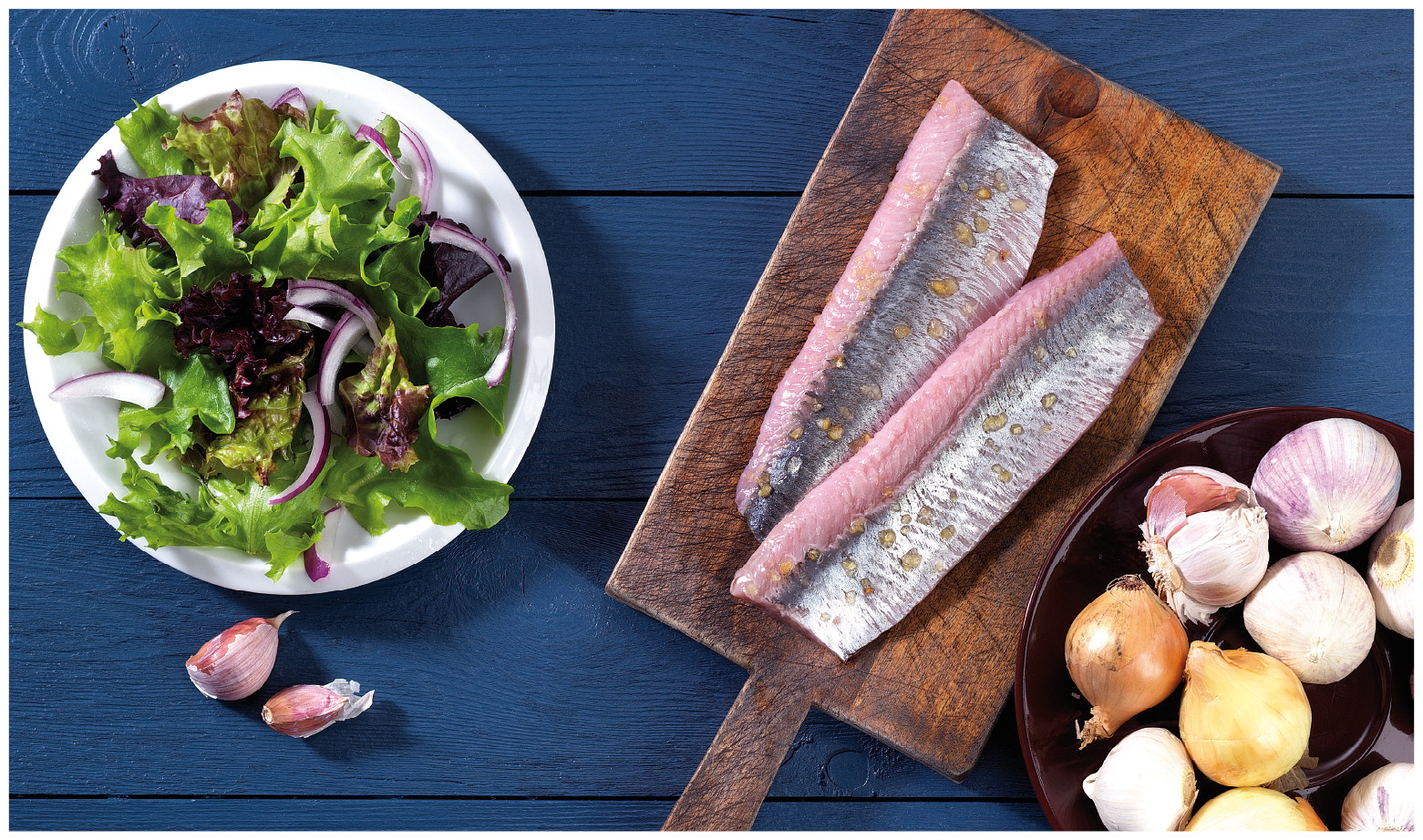 Zwei frische Heringsfilets auf einem Holzbrett, daneben ein Teller mit gemischtem Salat und Zwiebelringen, umgeben von Zwiebeln und Knoblauch auf einem blauen Tisch