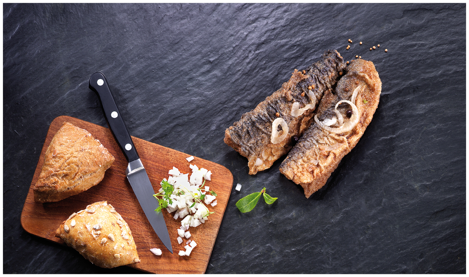 Gegrillter Fisch mit Zwiebeln und Brötchen auf Holzbrett Gegrillter Fisch mit Zwiebelringen, frischen Brötchen und Kräutern auf einem Holzbrett mit Messer, auf dunklem Untergrund