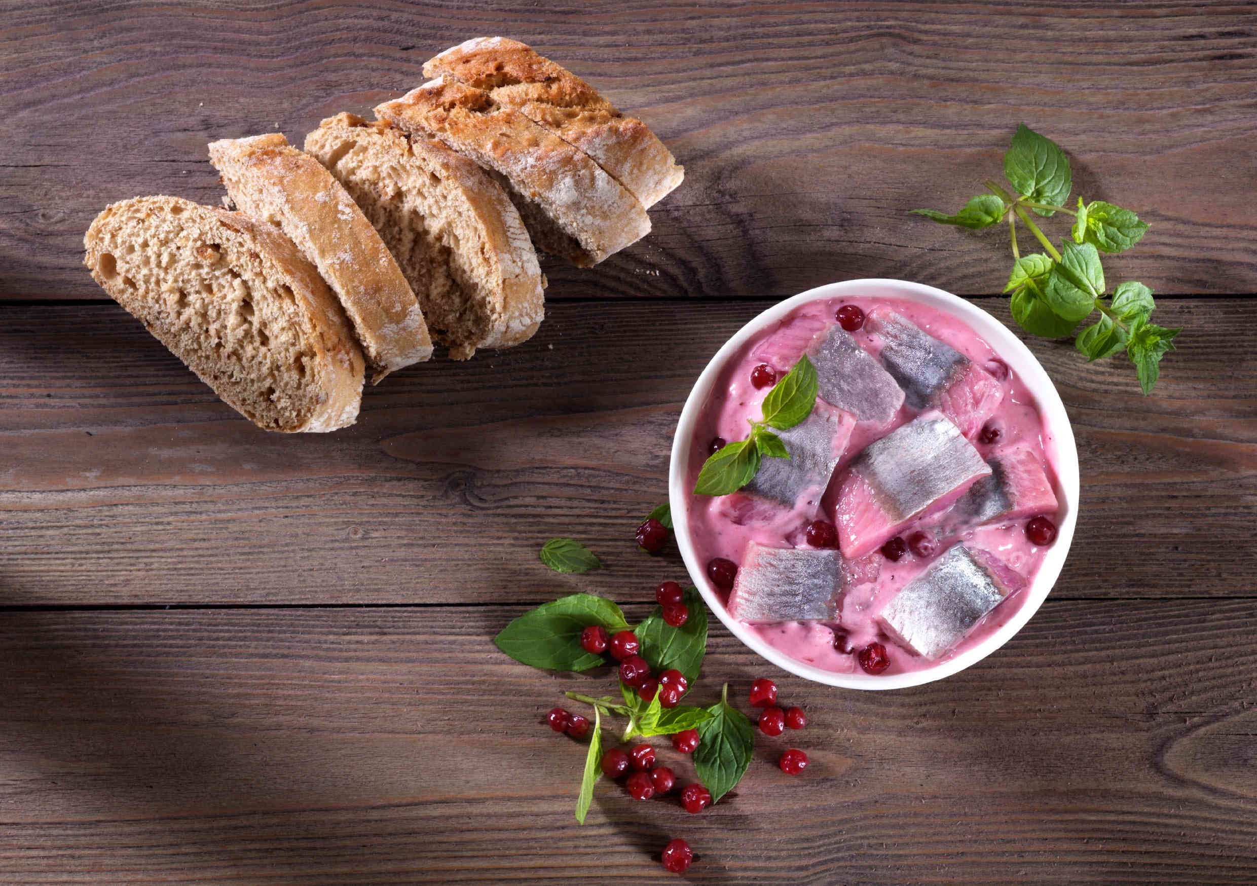 Eine Schüssel mit Heringssalat, verziert mit Preiselbeeren und frischen Kräutern, neben geschnittenem Brot auf einem Holztisch