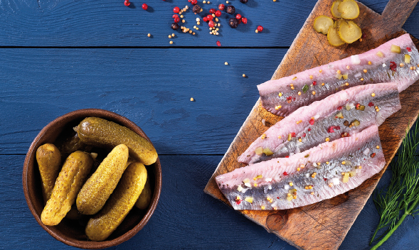 Heringsfilets mit Gewürzgurken und Gewürzen auf Holzbrett Drei hergerichtete Heringsfilets mit Gewürzen auf einem Holzbrett, daneben eine Schale gefüllt mit Gewürzgurken, fotografiert auf einem blauen Holztisch.