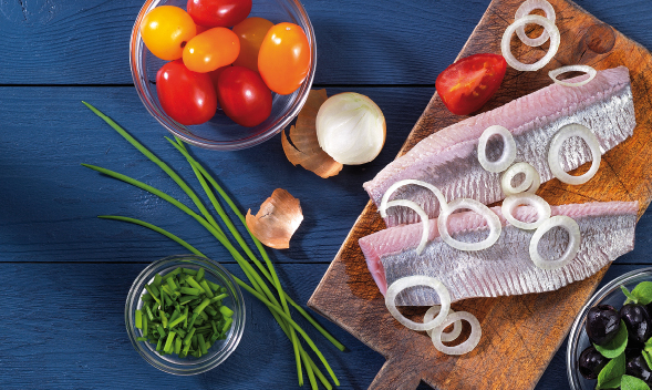 Ein hölzernes Schneidebrett mit Fischfilet, frischen Tomaten, Zwiebelringen und Schnittlauch auf einem blauen Holztisch