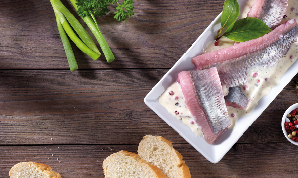 Heringsfilets in sahniger Soße mit Pfefferkörnern und Baguettescheiben Heringsfilets auf einem Teller in weißer Soße mit grünem Garnitur, Frühlingszwiebeln und Baguettescheiben auf einem Holztisch
