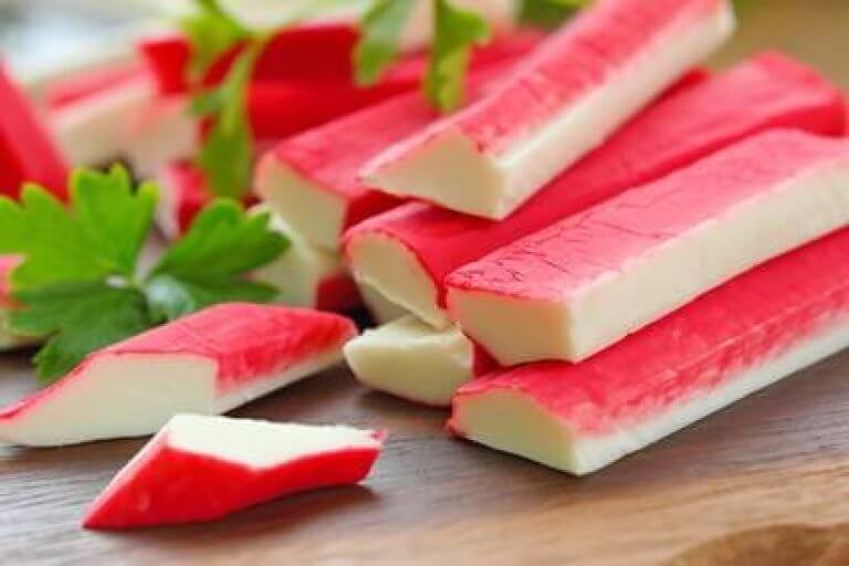 Frische Surimi-Sticks auf einem Holzbrett mit Petersilie Nahaufnahme von roten und weißen Surimi-Sticks, dekoriert mit grünen Petersilienblättern auf einem Holzbrett