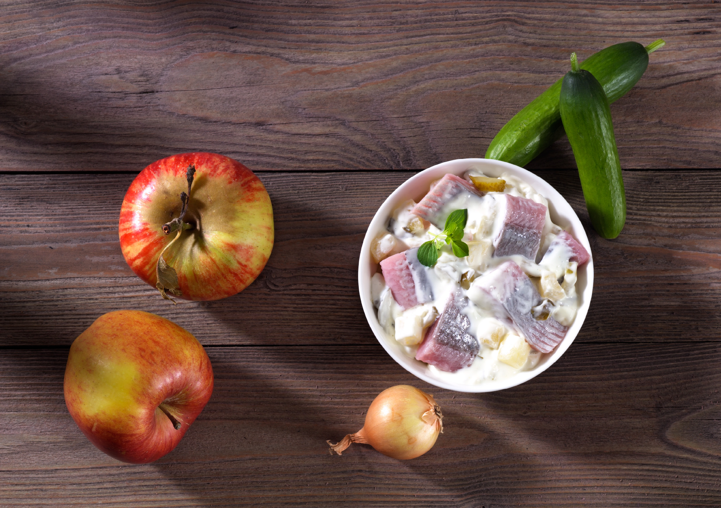 Schüssel mit Heringssalat, umgeben von zwei Äpfeln, einer Zwiebel und zwei Gurken auf einem Holztisch