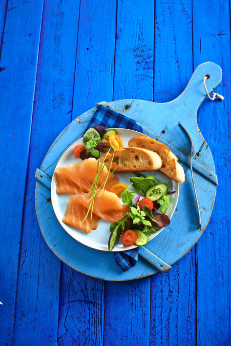Frischer Lachs mit Salat und Brot auf blauem Holzbrett Ein Teller mit Räucherlachs, Salat und Brot auf einem rustikalen blauen Holzbrett angerichtet