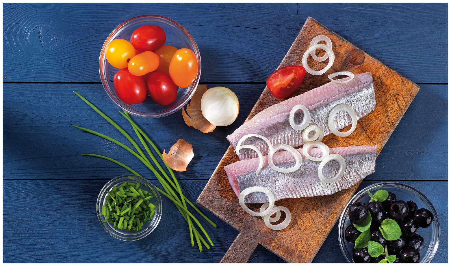 Frische Heringfilets auf Holzbrett mit Zwiebelscheiben, daneben Tomaten, Schnittlauch und Oliven.