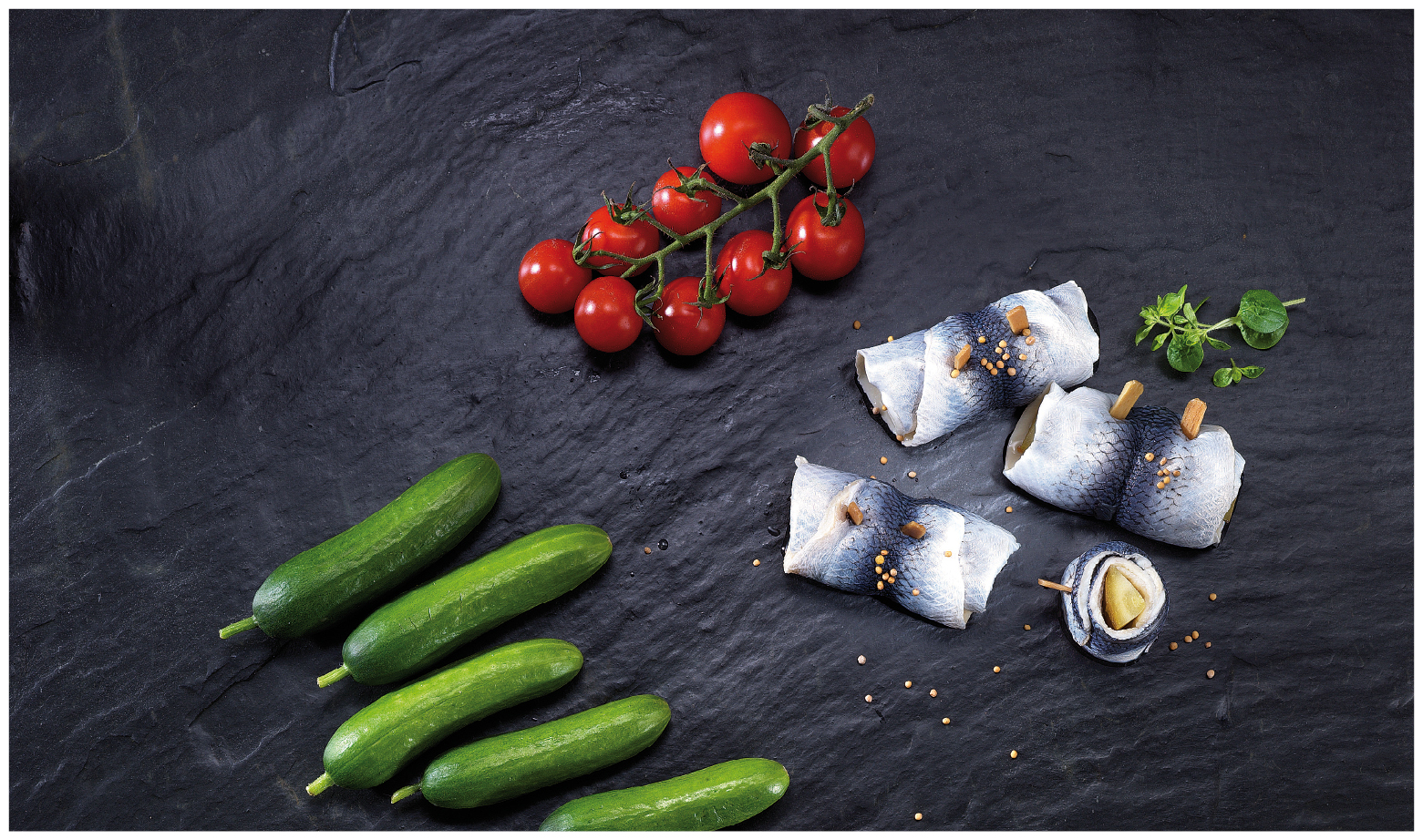 Frische Zutaten: Tomaten, Gurken und Rollmops auf Schieferplatte Auf einer Schieferplatte liegen frische Tomaten, Gurken und Rollmops, garniert mit Kräutern und Senfkörnern.