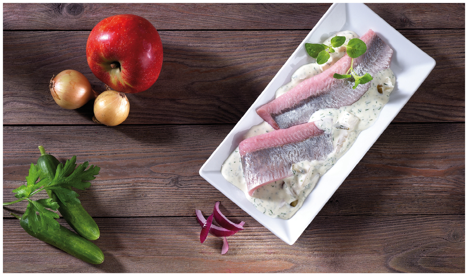 Matjesfilets in Sahnesauce mit Apfel, Zwiebeln und Gurke Frische Matjesfilets in cremiger Sahnesauce auf einem weißen Teller, umgeben von Apfel, Zwiebeln und Gurken auf einem Holztisch