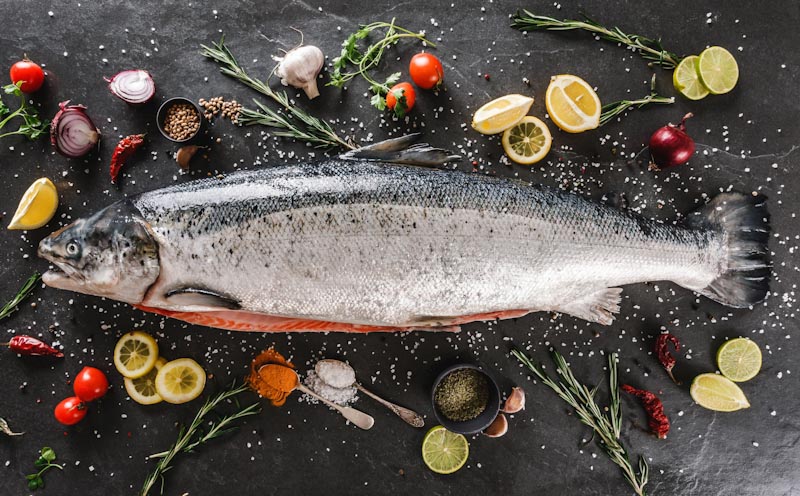 Frischer Lachs auf Tisch mit Zitrone, Kräutern und Gewürzen Ein ganzer frischer Lachs liegt auf einem Tisch, umgeben von Zitronenscheiben, Kräutern wie Rosmarin und Gewürzen, darunter Salz, Pfeffer und Knoblauch.