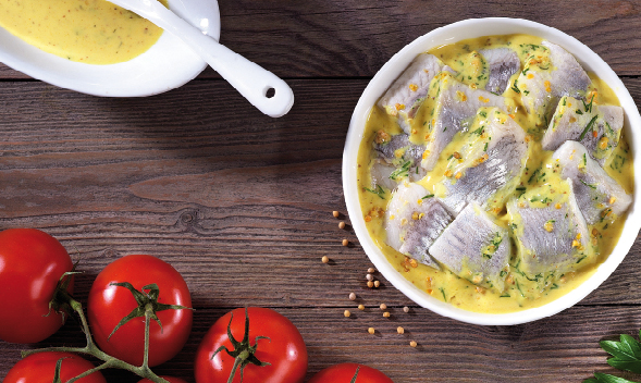 Weiße Schale mit Heringsfilet in Senf-Dill-Sauce auf Holztisch, daneben frische Tomaten