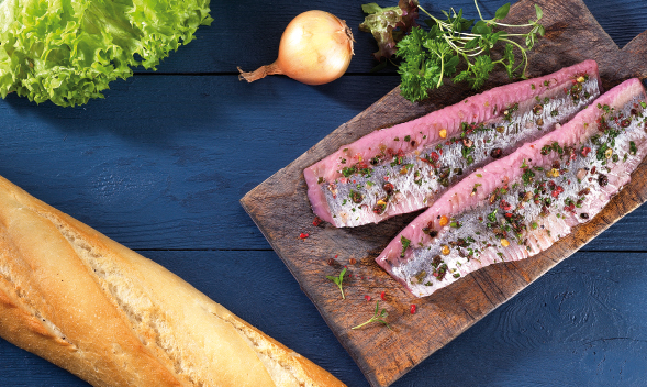 Frische Fischfilets mit Gewürzen auf Holzbrett Fischfilets mit Kräutern auf Schneidebrett, daneben Baguette, Salat, Zwiebel und Petersilie auf dunklem Hintergrund.