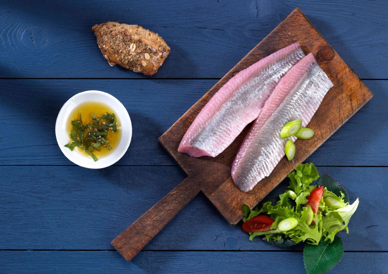 Frische Fischfilets auf Holzbrett mit Salat und Baguette Zwei frische Fischfilets auf einem hölzernen Schneidebrett, begleitet von frischem Salat und einem Stück Baguette auf einem blauen Hintergrund