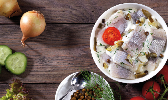 Traditioneller Matjessalat mit Zwiebeln und Tomaten Schüssel mit Matjessalat, garniert mit Tomaten und Zwiebeln, daneben Gurkenscheiben und Gewürze auf Holztisch