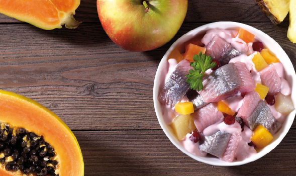 Schüssel mit Heringssalat, umgeben von Papaya, Apfel und Ananas auf einem Holztisch